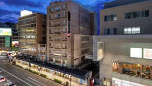 Un hotel de varios pisos con un diseño moderno, situado en una esquina con entrada visible y un área exterior techada.
