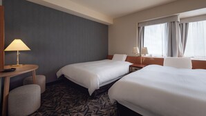Habitación de hotel con dos camas, una mesita de noche con lámpara, una silla y un ventanal con cortinas.