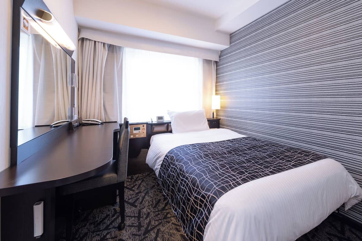 A hotel room with a bed, a desk, a chair, a nightstand, a lamp, and a window with curtains.