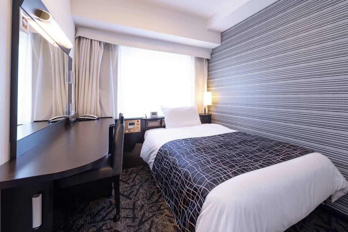 Habitación de hotel con cama, escritorio, silla, mesita de noche, lámpara y ventana con cortinas.