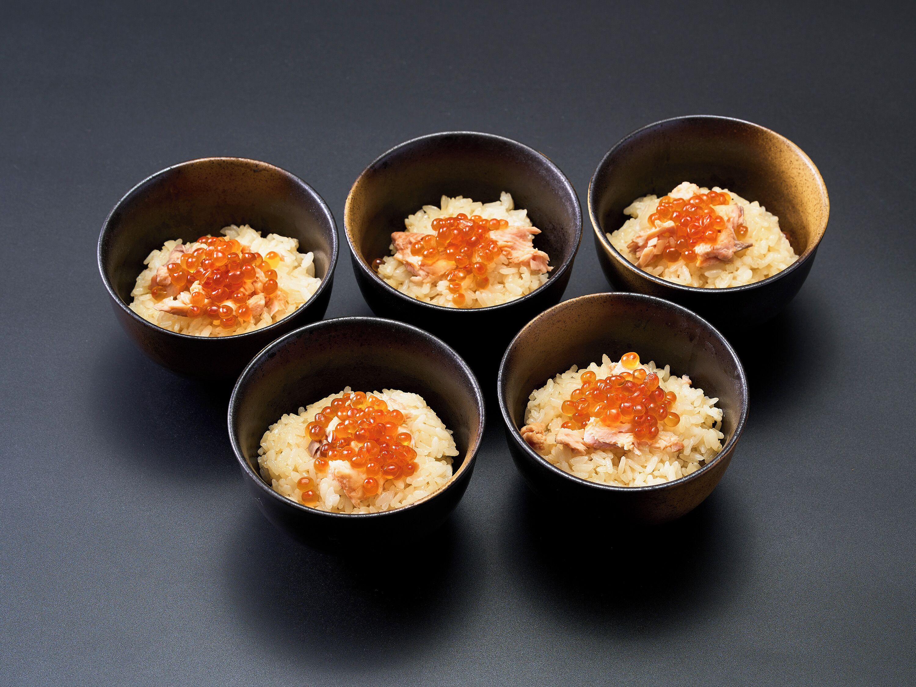 Five bowls of rice with fish and roe on a dark surface.