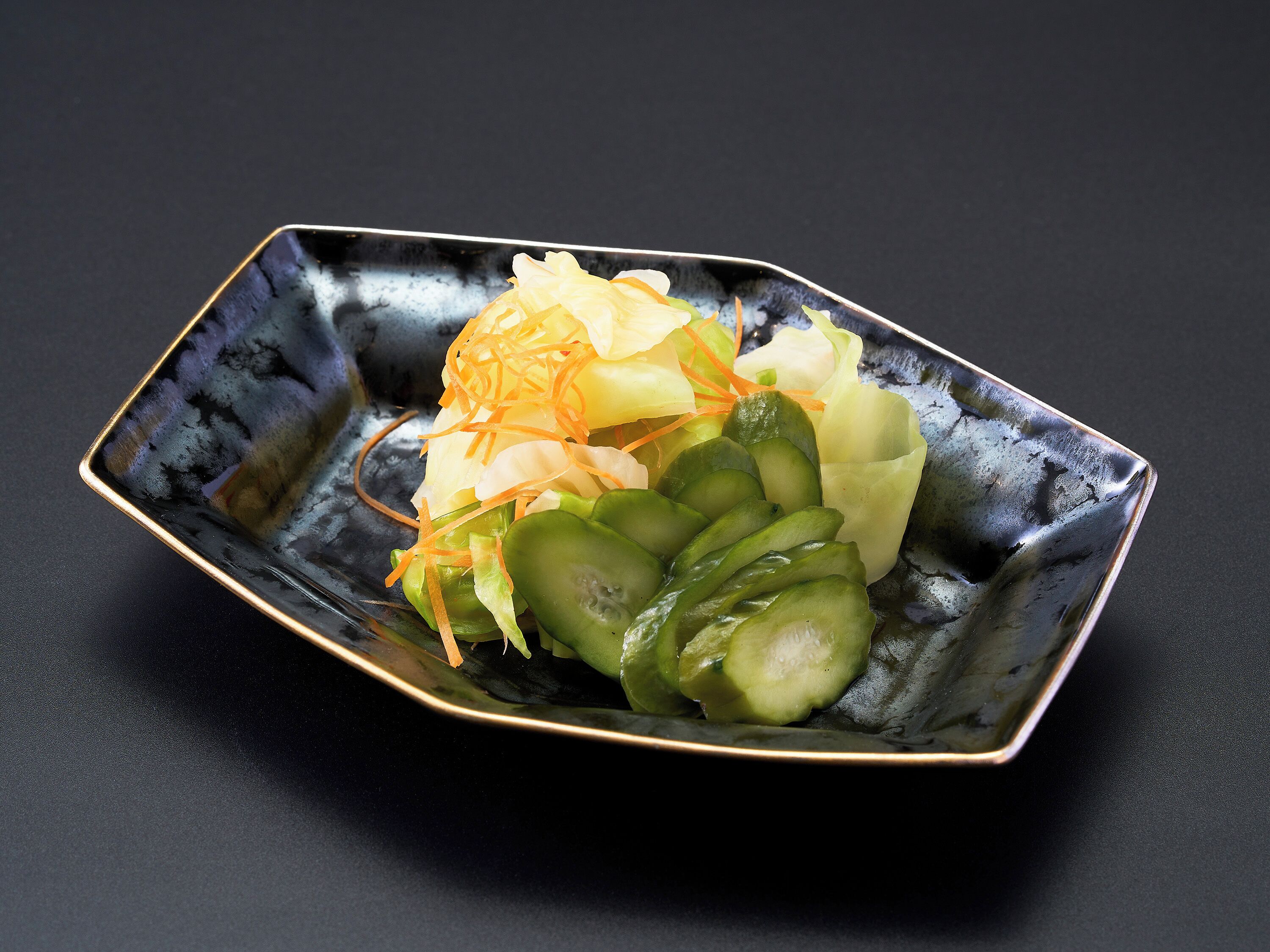 A rectangular black ceramic dish with a gold rim, containing sliced cucumbers, shredded carrots, and a translucent vegetable.