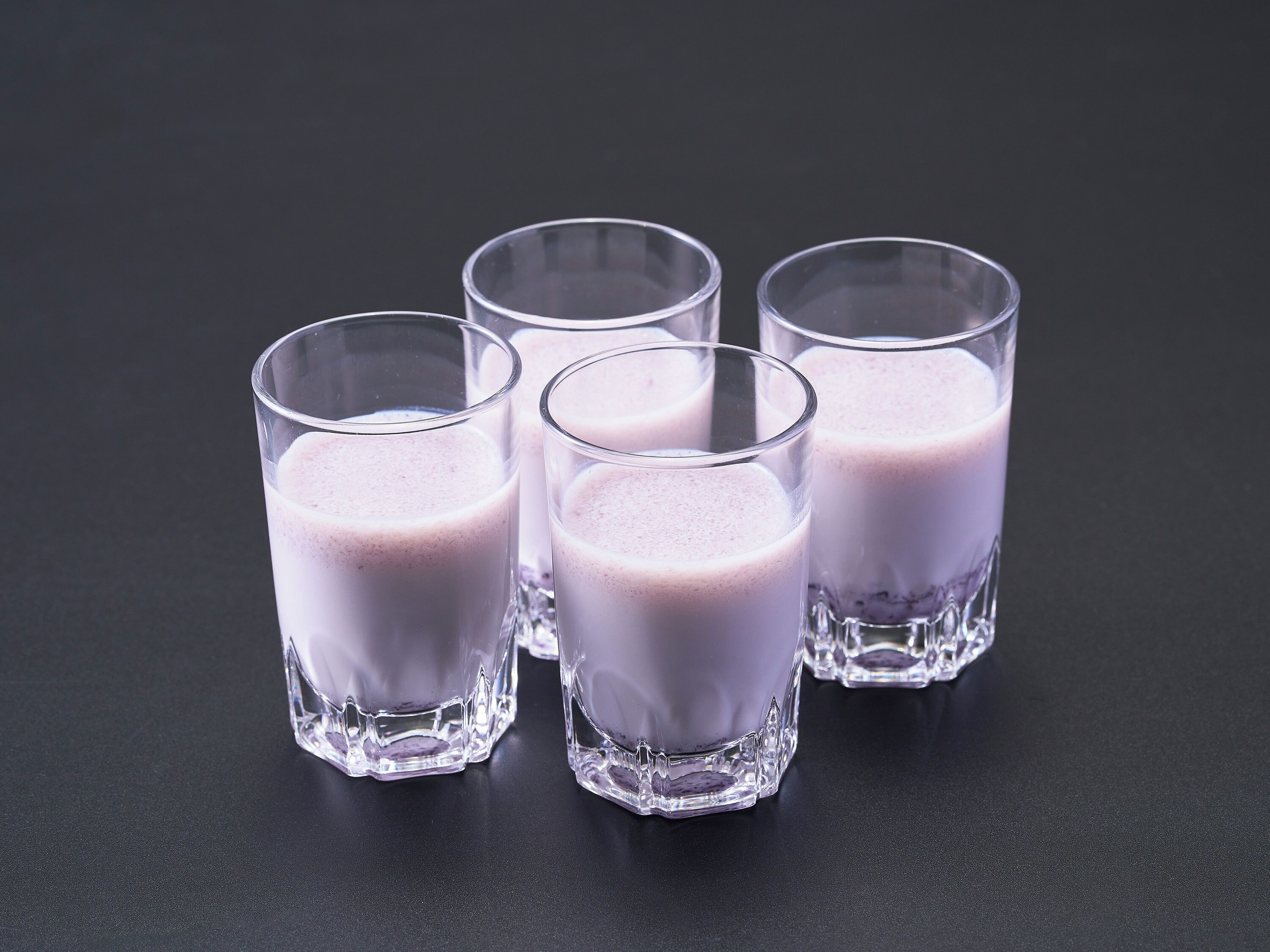 Four clear glass cups with a purple beverage on a dark surface.