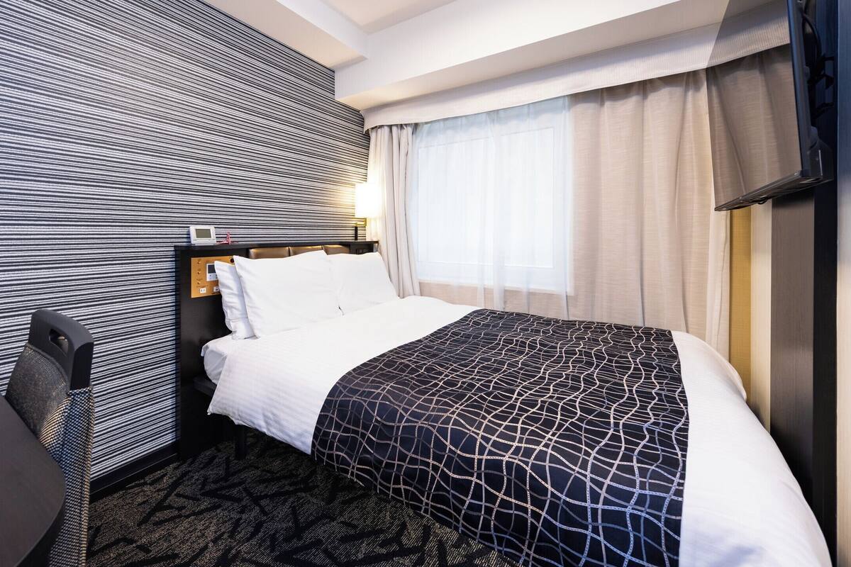 A hotel room with a bed, a desk, a chair, a control panel, and a television.