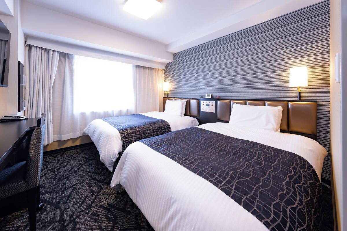 A hotel room with two beds, a desk, a television, and a control panel.