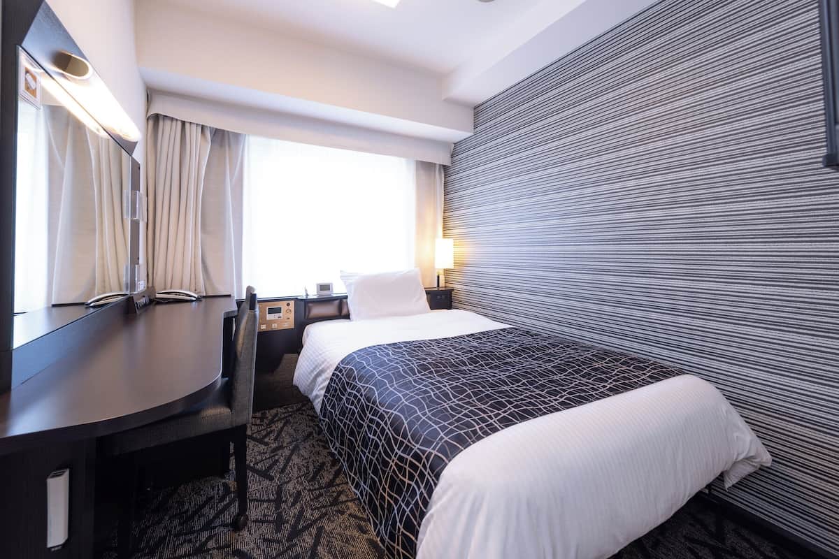 A hotel room with a bed, a desk, a chair, a large mirror, a window with curtains, and a wall with a textured pattern.