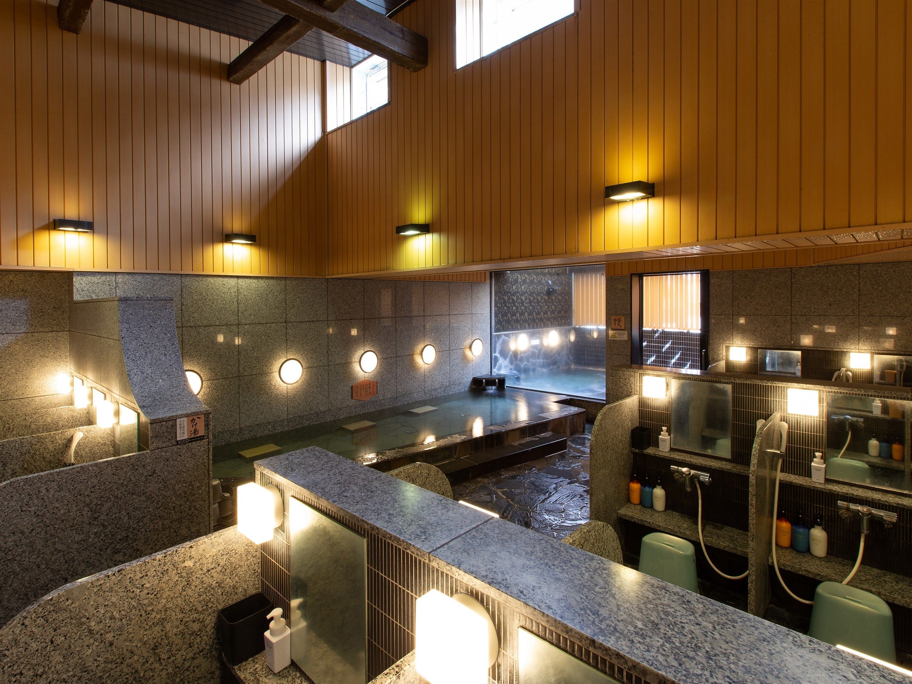 A traditional Japanese onsen with a stone hot spring, wooden paneling, and a counter with amenities.