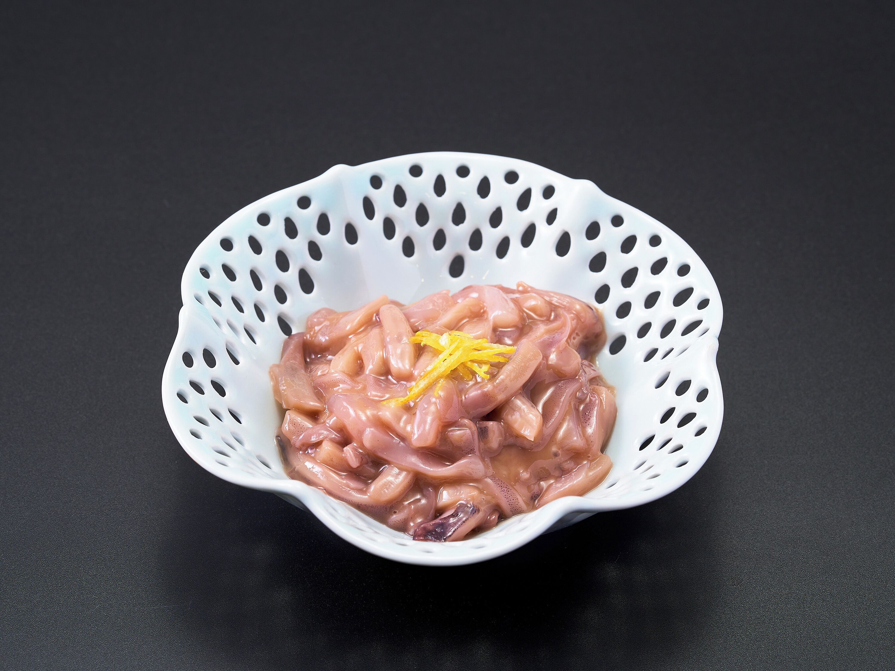 A white colander containing raw chicken pieces with a yellow zest garnish on a black surface.