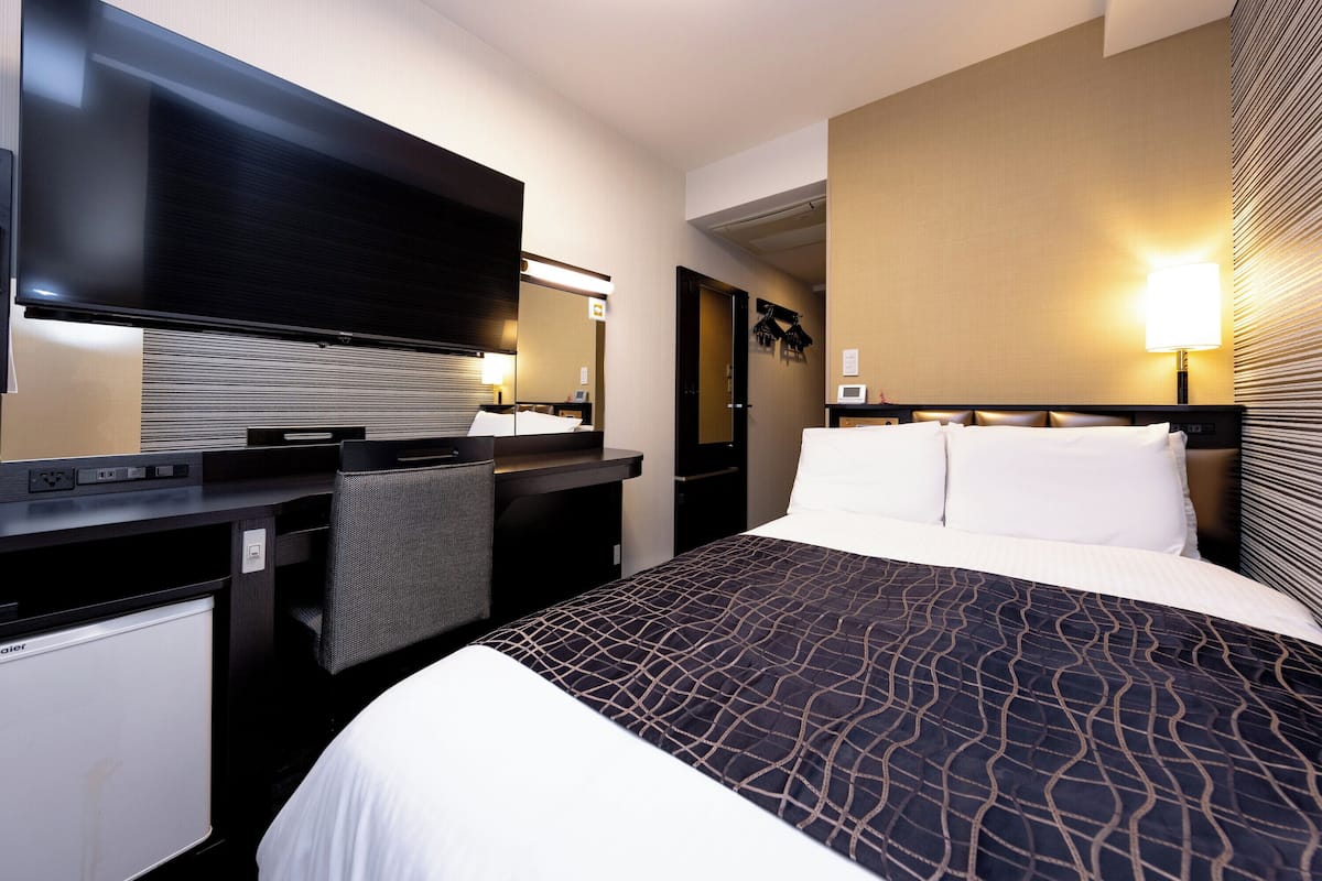 A hotel room with a bed, a desk, a flat-screen TV, a mirror, and a wall-mounted lamp.