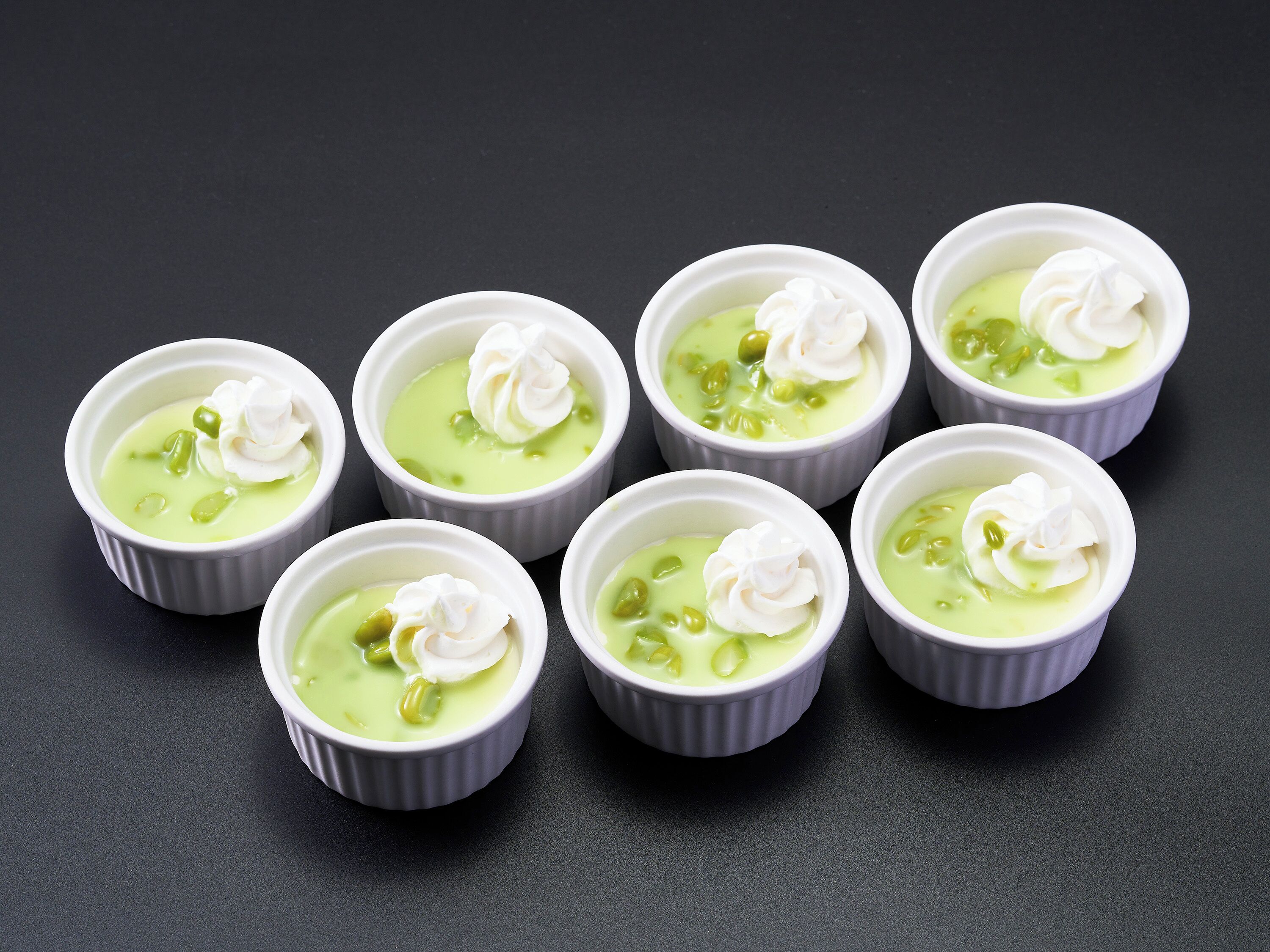 Sei piccoli recipienti bianchi con dessert verde e panna montata su una superficie scura.