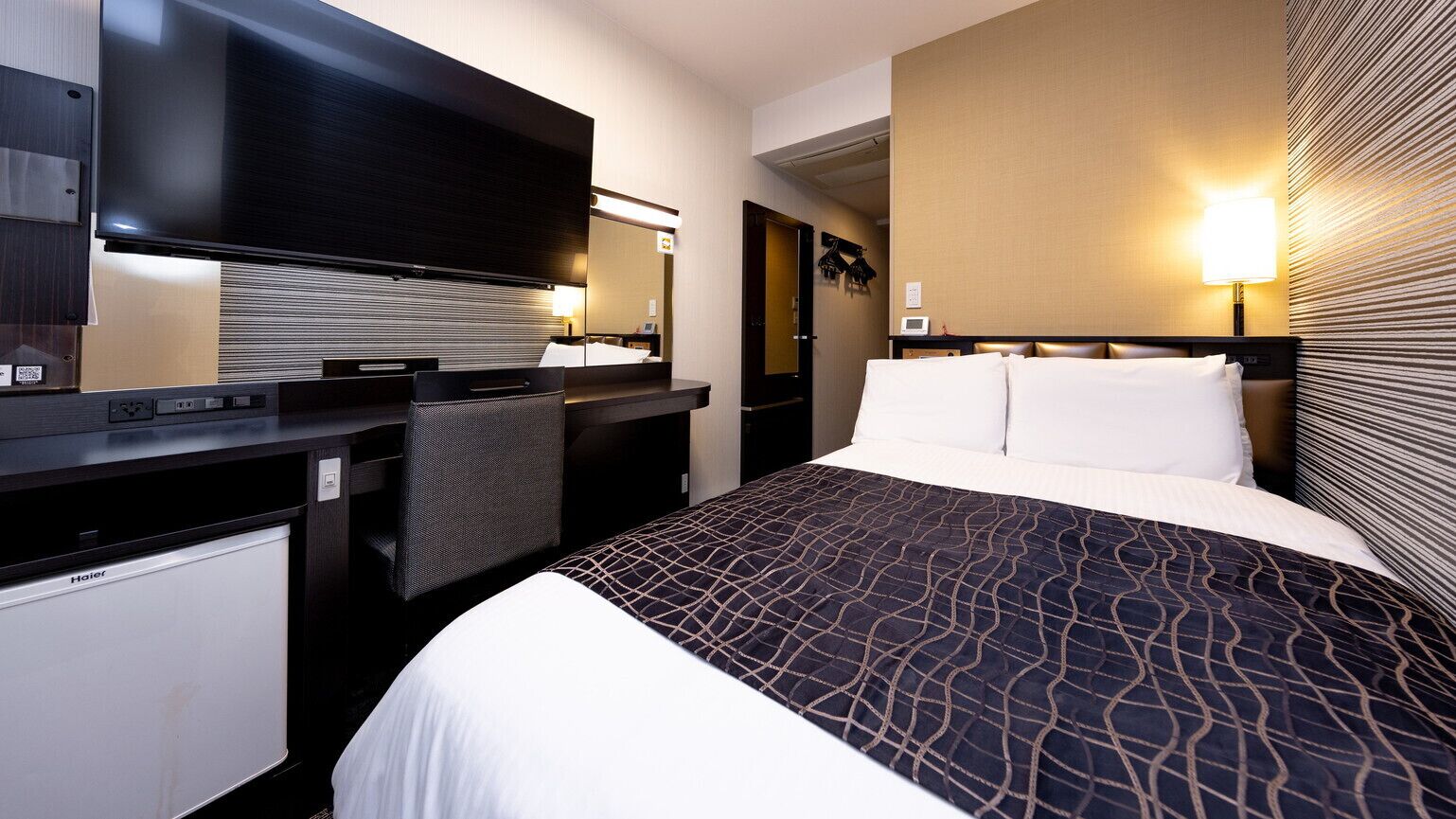 Une chambre d’hôtel équipée d’un lit, d’un bureau avec un téléviseur à écran plat, d’un mini-réfrigérateur et d’une lampe fixée au mur.