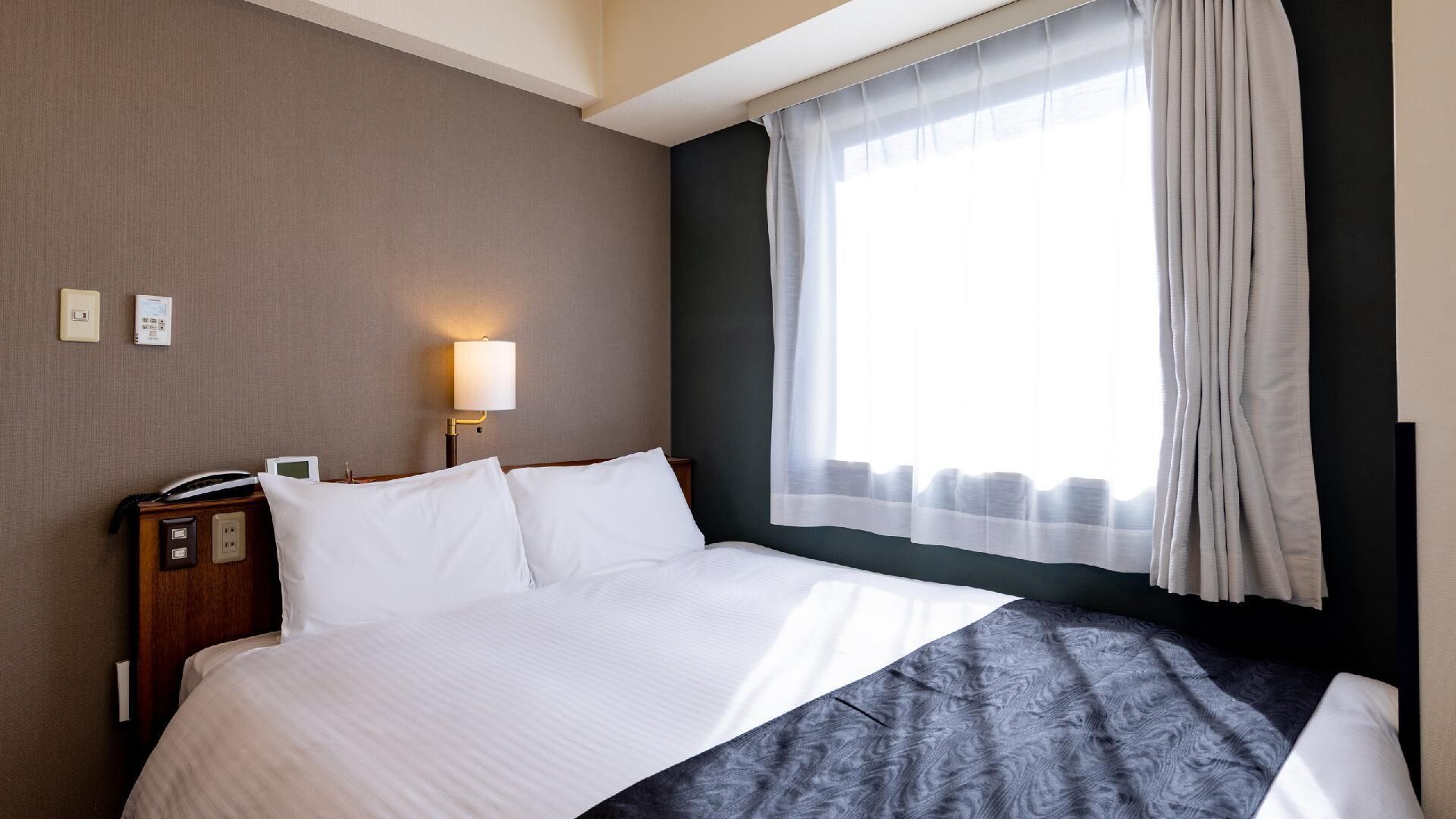 A hotel room with a bed, a bedside lamp, a telephone, and a window with curtains.