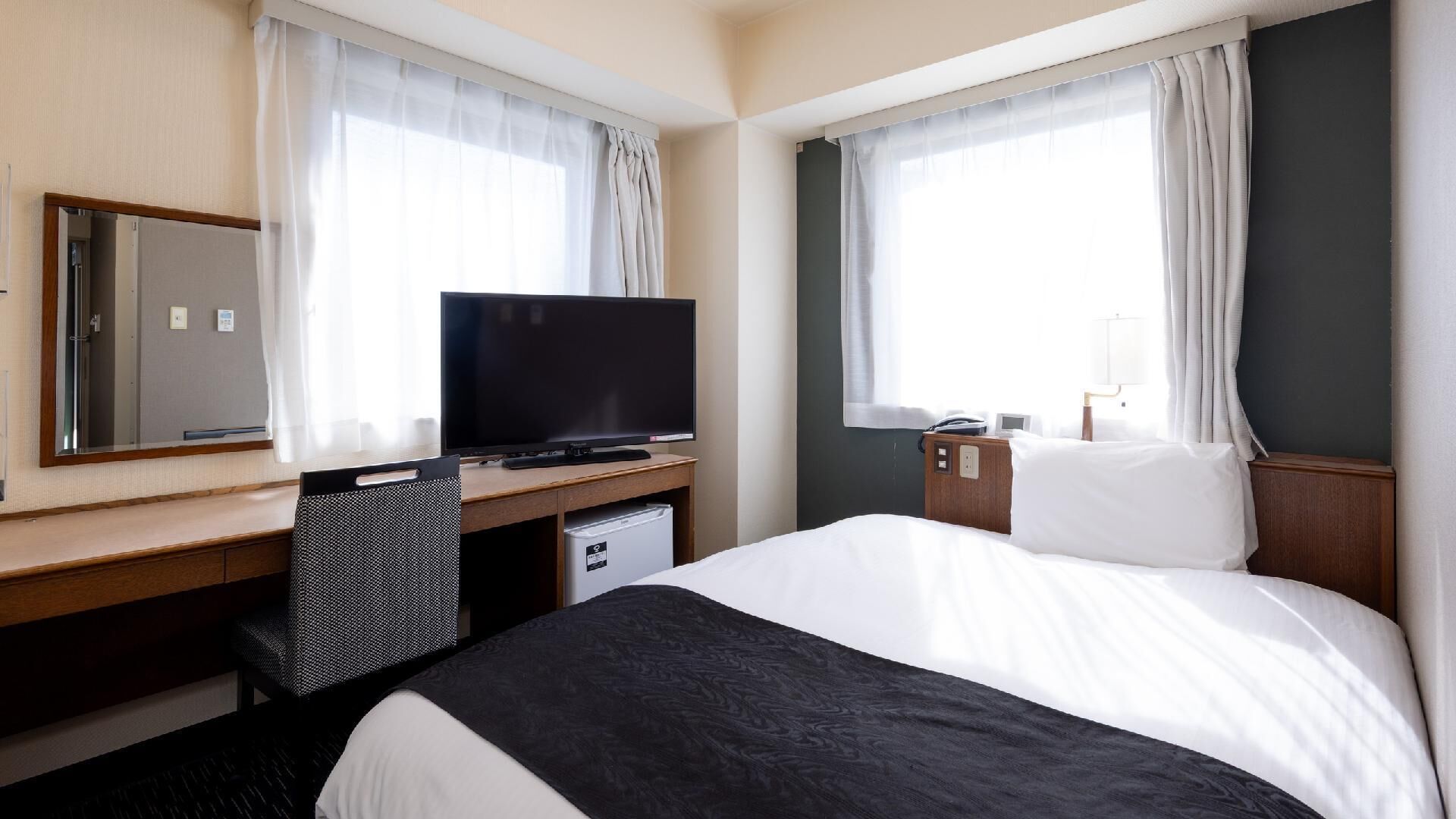 A hotel room with a bed, a desk with a television, a chair, a small desk, and a mirror.