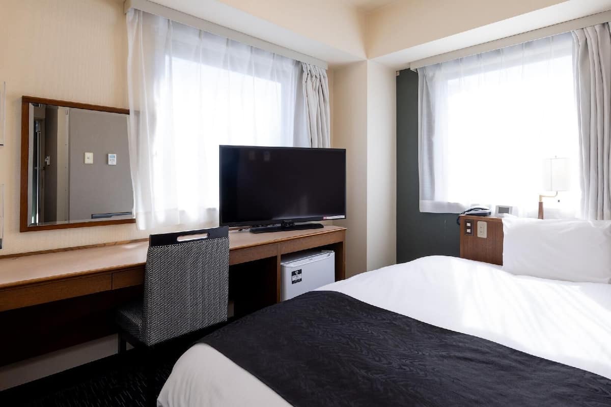 A hotel room with a bed, a desk with a television, a chair, a small desk, and a mirror.