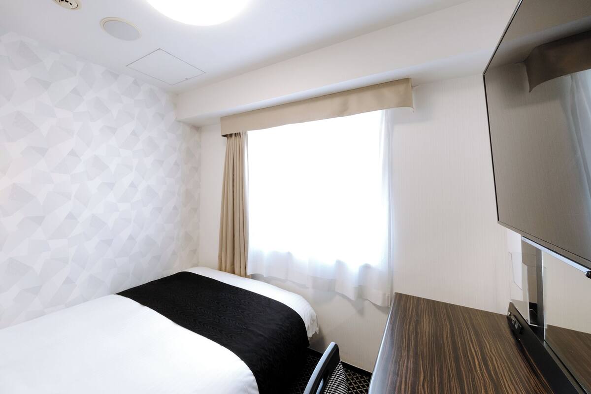 Habitació d'hotel amb llit, televisió, escriptori i finestra amb cortines.