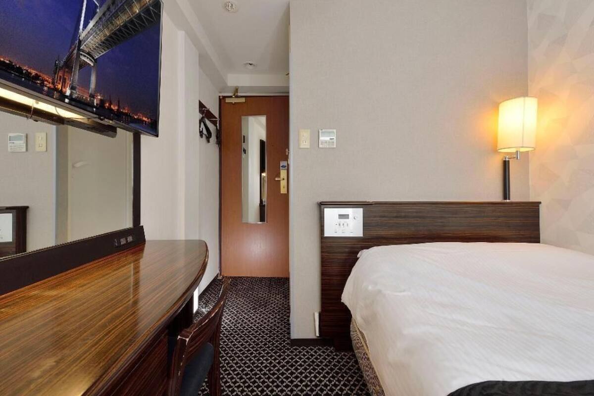 Una habitació d'hotel amb un llit, escriptori, televisió penjada a la paret i làmpada.