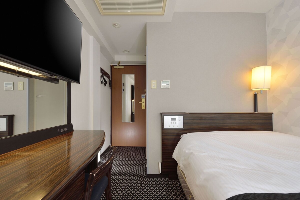 A hotel room with a bed, a desk, a television, a wall-mounted lamp, and a door leading to another room.