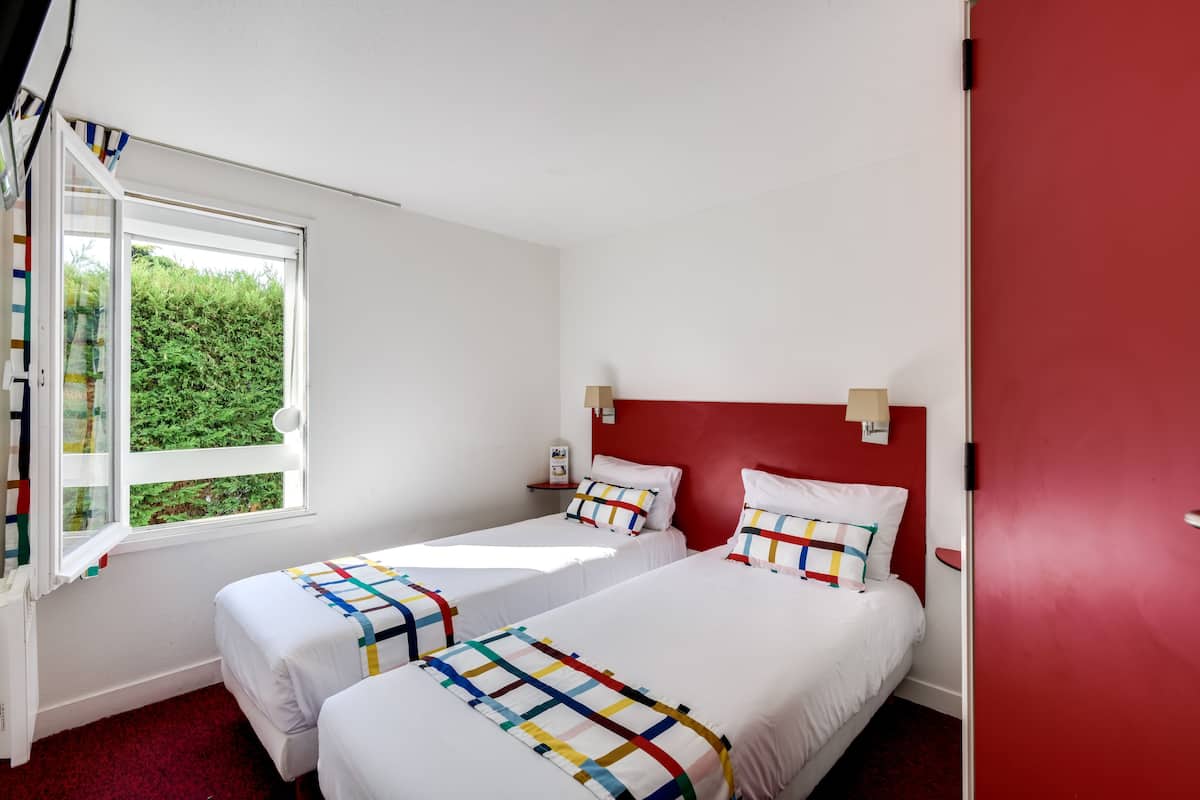 Een hotelkamer met twee eenpersoonsbedden, een rode accentmuur, een raam met uitzicht op groen, en een wit plafond.