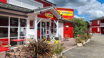 Een rood-wit gebouw met een bord waarop “balladins initial” en “RECEPTION” staat. Er zijn buitentafels en stoelen, en potplanten.