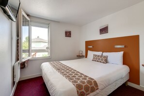 Een hotelkamer met een groot bed, een raam met uitzicht, een thermostaat en een kleine tafel met een lamp.