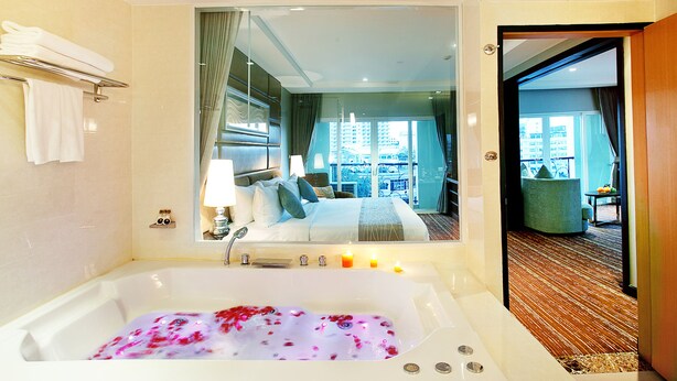 A hotel room with a large bathtub filled with water and rose petals, a bed with white linens, a nightstand with a lamp, and a view of the city through large windows. - Intimate Hotel Pattaya (Pattaya)