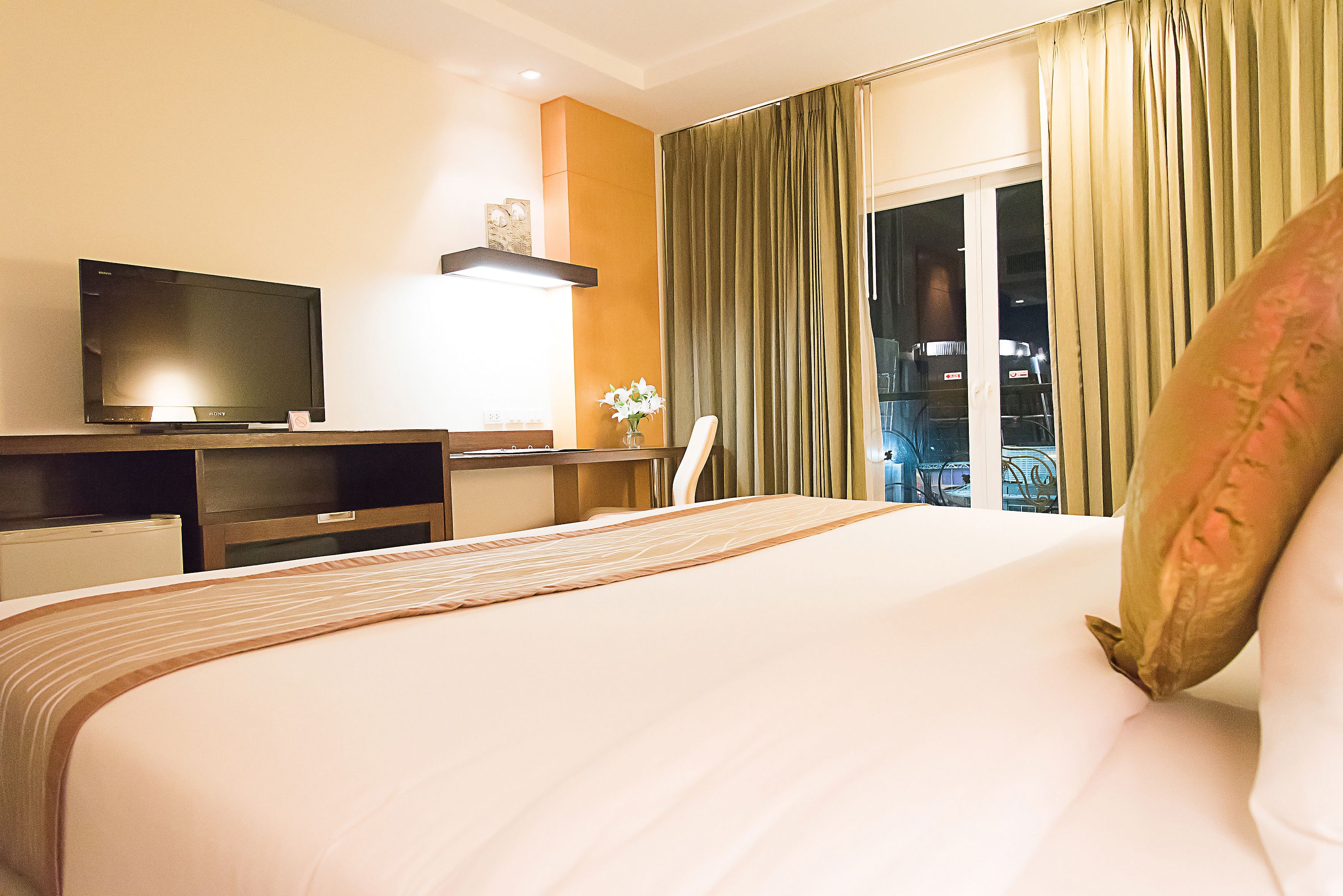 A hotel room with a bed, a desk with a flat-screen TV, a chair, and a window with curtains.