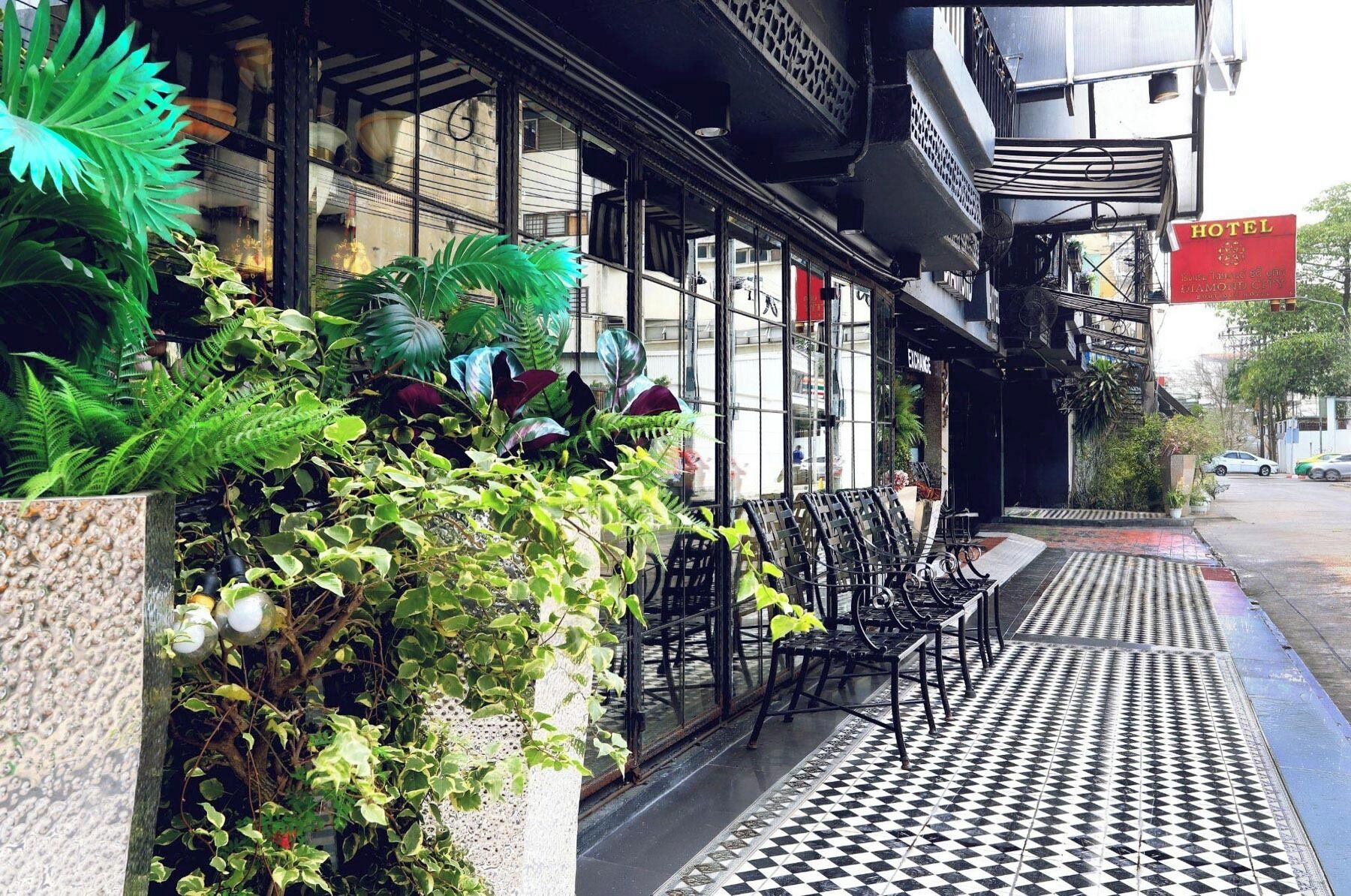 Entrada de hotel con suelo a cuadros, mesas al aire libre y abundante vegetación.