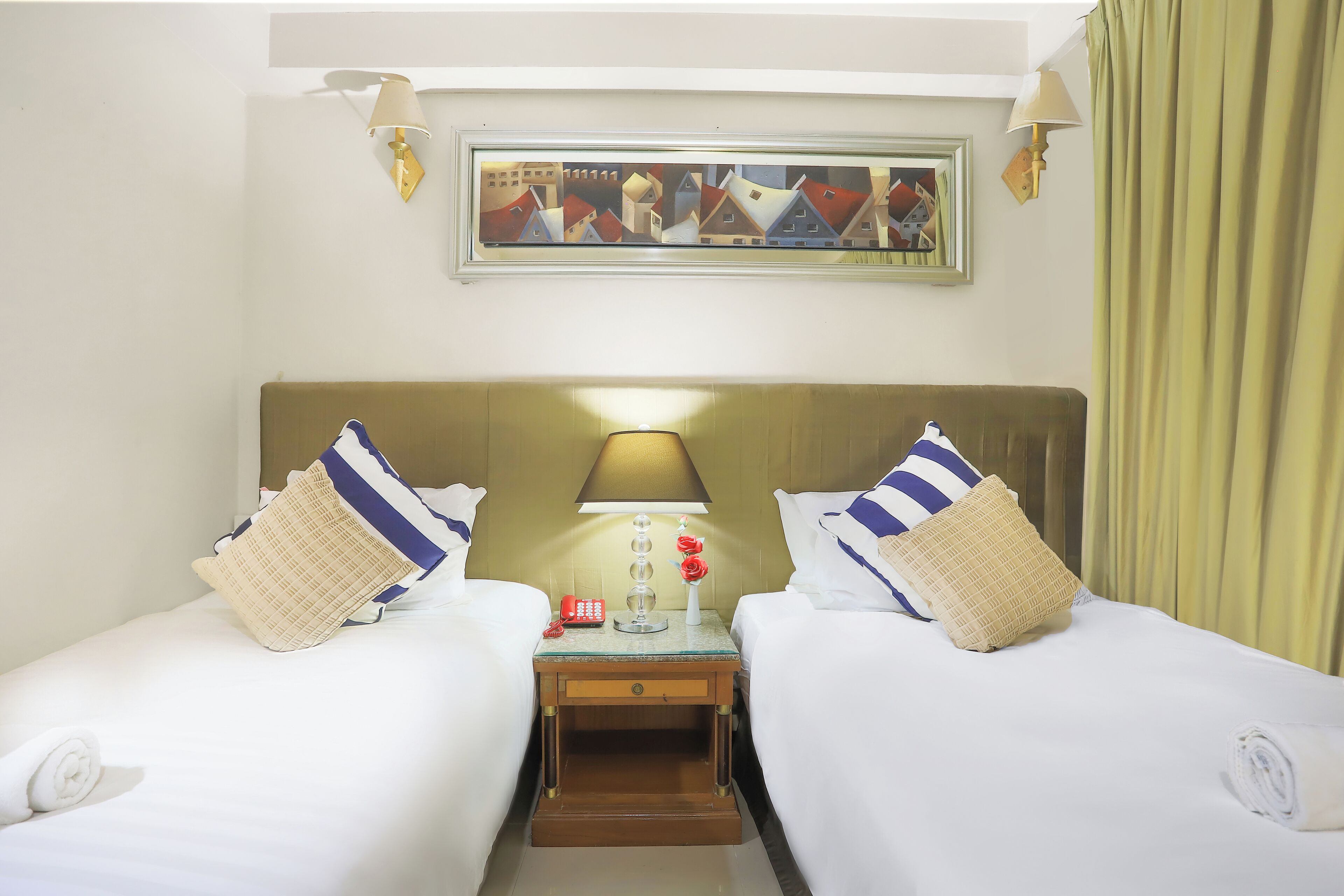 A hotel room with two beds, a nightstand, a lamp, a telephone, and a painting on the wall.