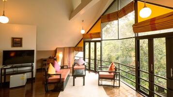 Relax in a spacious living area with a high ceiling and large windows offering a view of the surrounding greenery. Enjoy a flat-screen TV, refrigerator, and comfortable seating area.