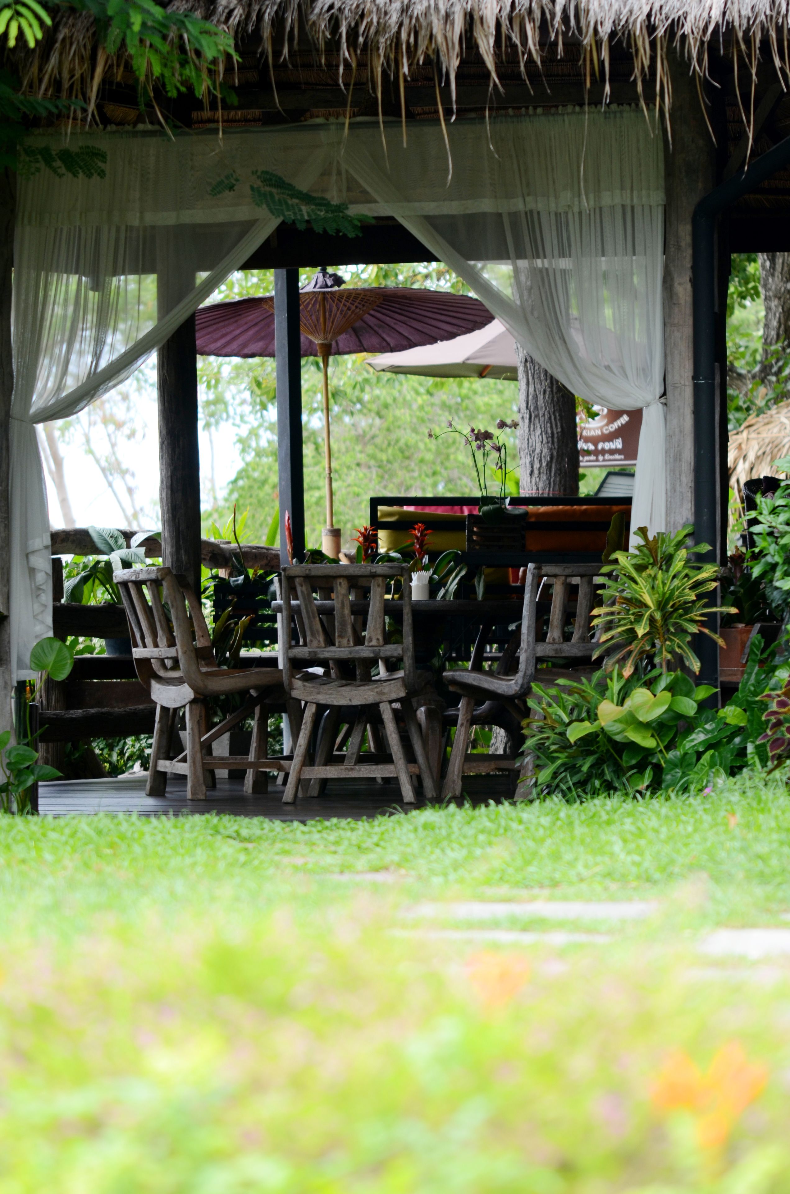 A wooden outdoor seating area with chairs and a table, surrounded by lush greenery and a thatched roof structure.