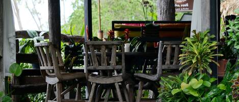 A wooden outdoor seating area with chairs and a table, surrounded by lush greenery and a thatched roof structure.