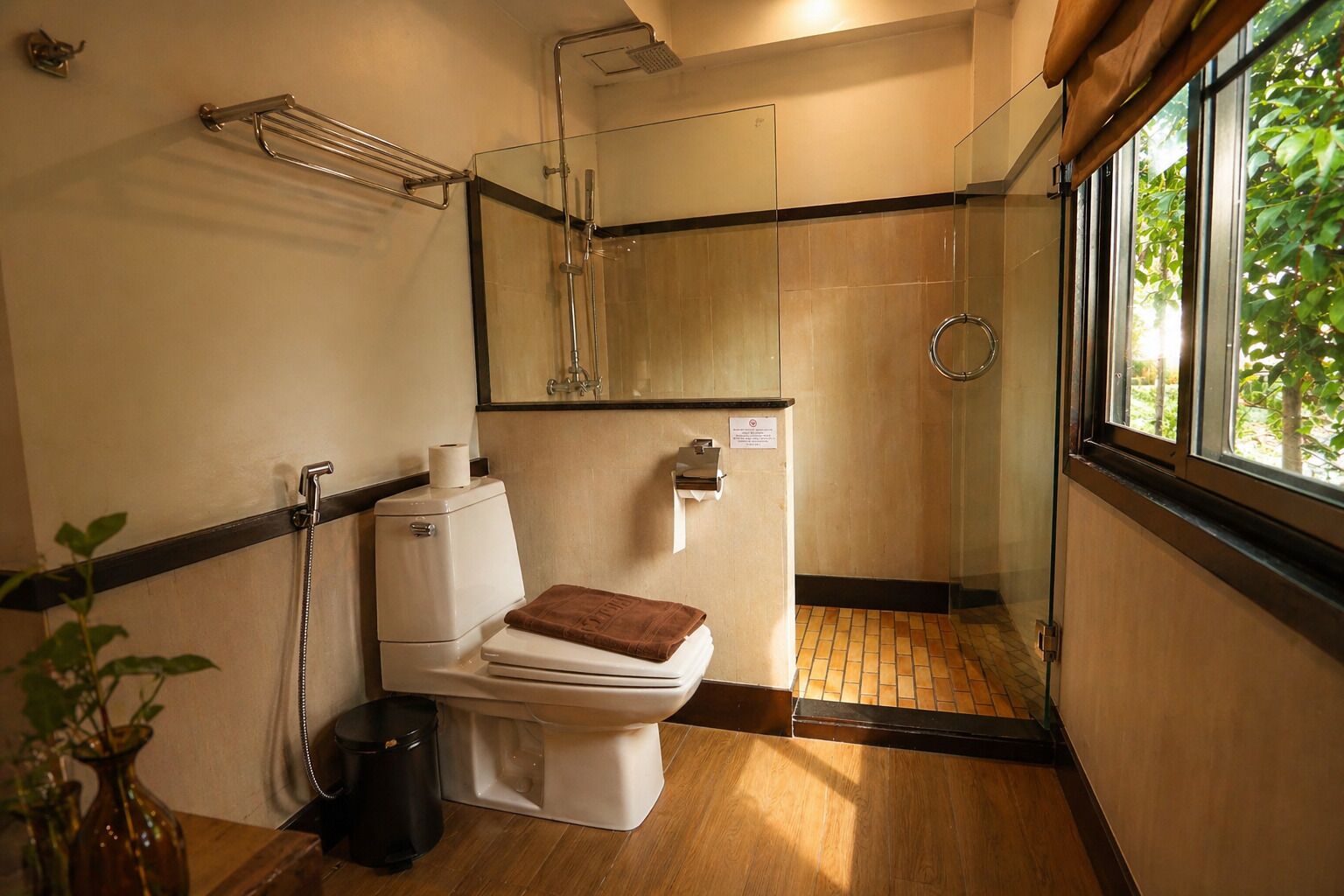 A compact bathroom with a toilet, a shower, a glass partition, and a window with a view of greenery.
