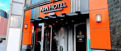 Exterior del APA HOTEL con un diseño moderno, destacando una fachada naranja prominente y una entrada amplia.