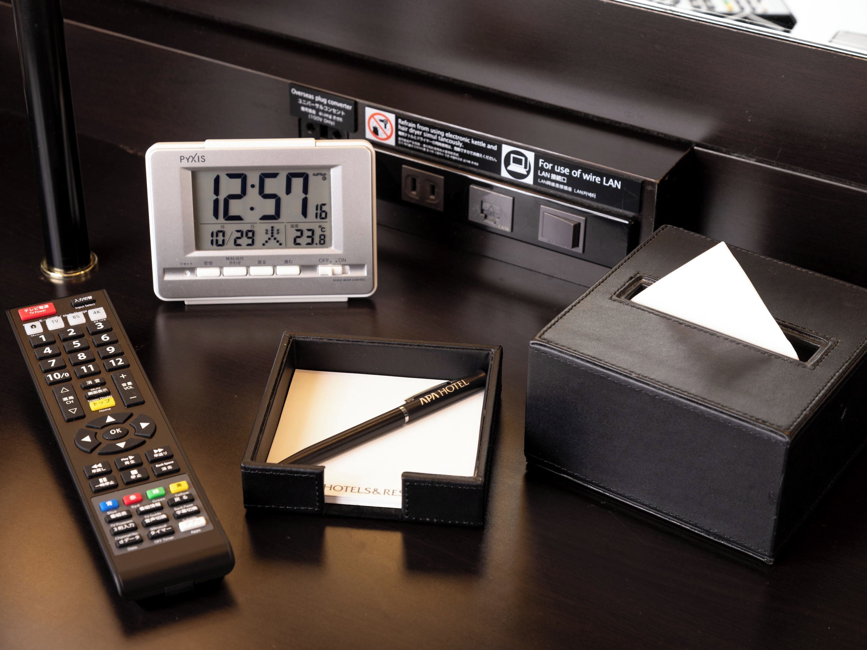Un bureau d’hôtel avec une horloge numérique, une télécommande, un stylo et une boîte de mouchoirs.