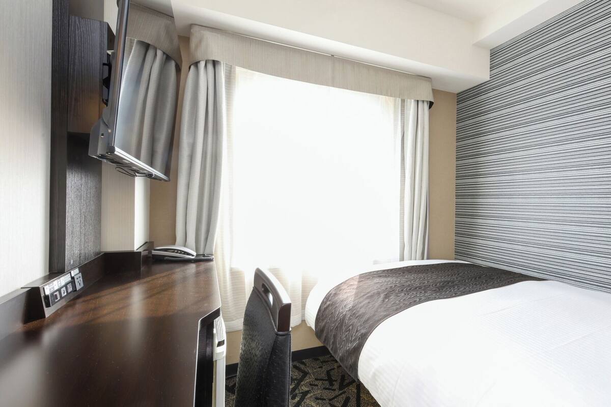 A hotel room with a bed, desk, chair, and a large window with curtains.