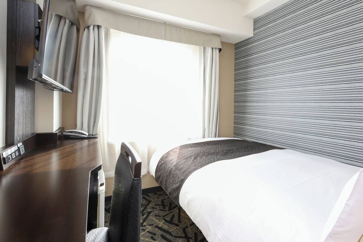 A hotel room with a desk, chair, bed, and a large window with curtains.