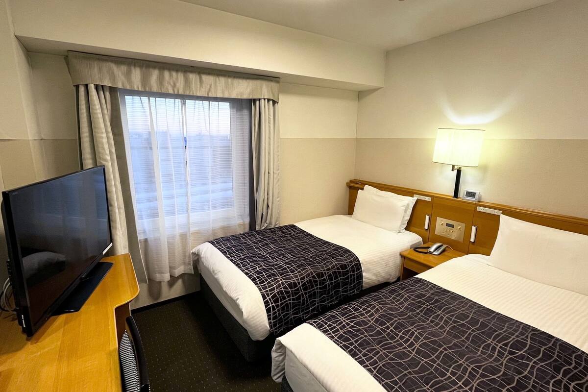 A hotel room with two beds, a TV, a bedside table, a lamp, and a window with curtains.