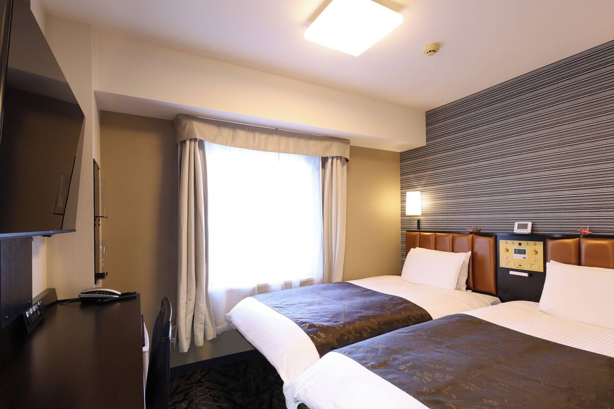 A hotel room with two beds, a flat-screen TV, a control panel on the wall, and a window with curtains.