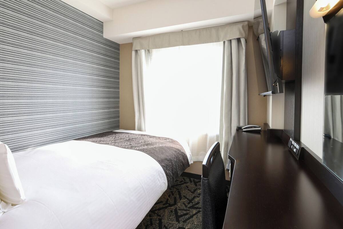 A hotel room with a bed, a desk, a television, and a window with curtains.