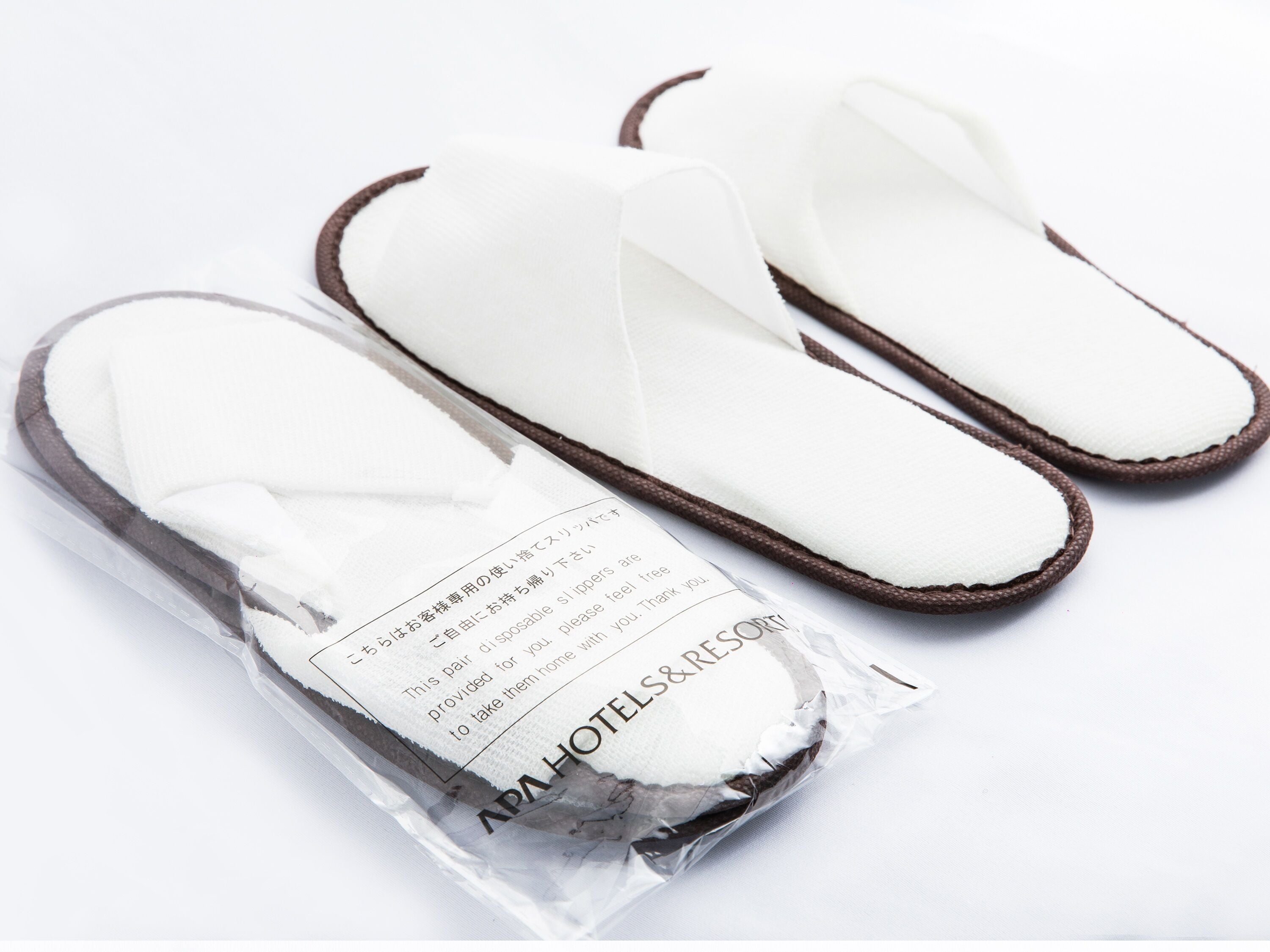 A pair of white slippers with brown trim, packaged in a clear plastic bag with a label.
