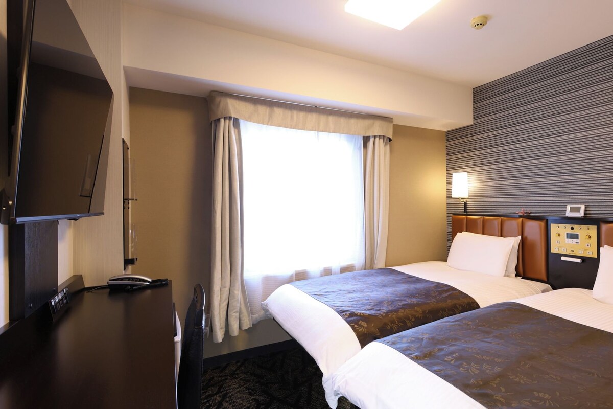 Ein Hotelzimmer mit zwei Betten, einem Flachbildfernseher, einem Nachttisch und einer Bedienfeldtafel an der Wand.
