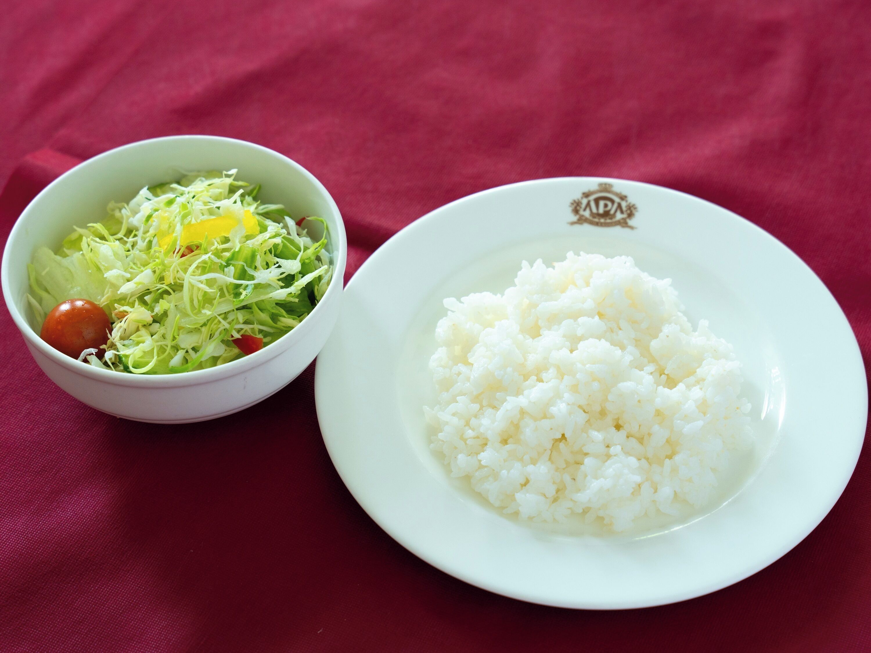 Un tazón blanco con ensalada y un plato de arroz blanco sobre un mantel rojo.