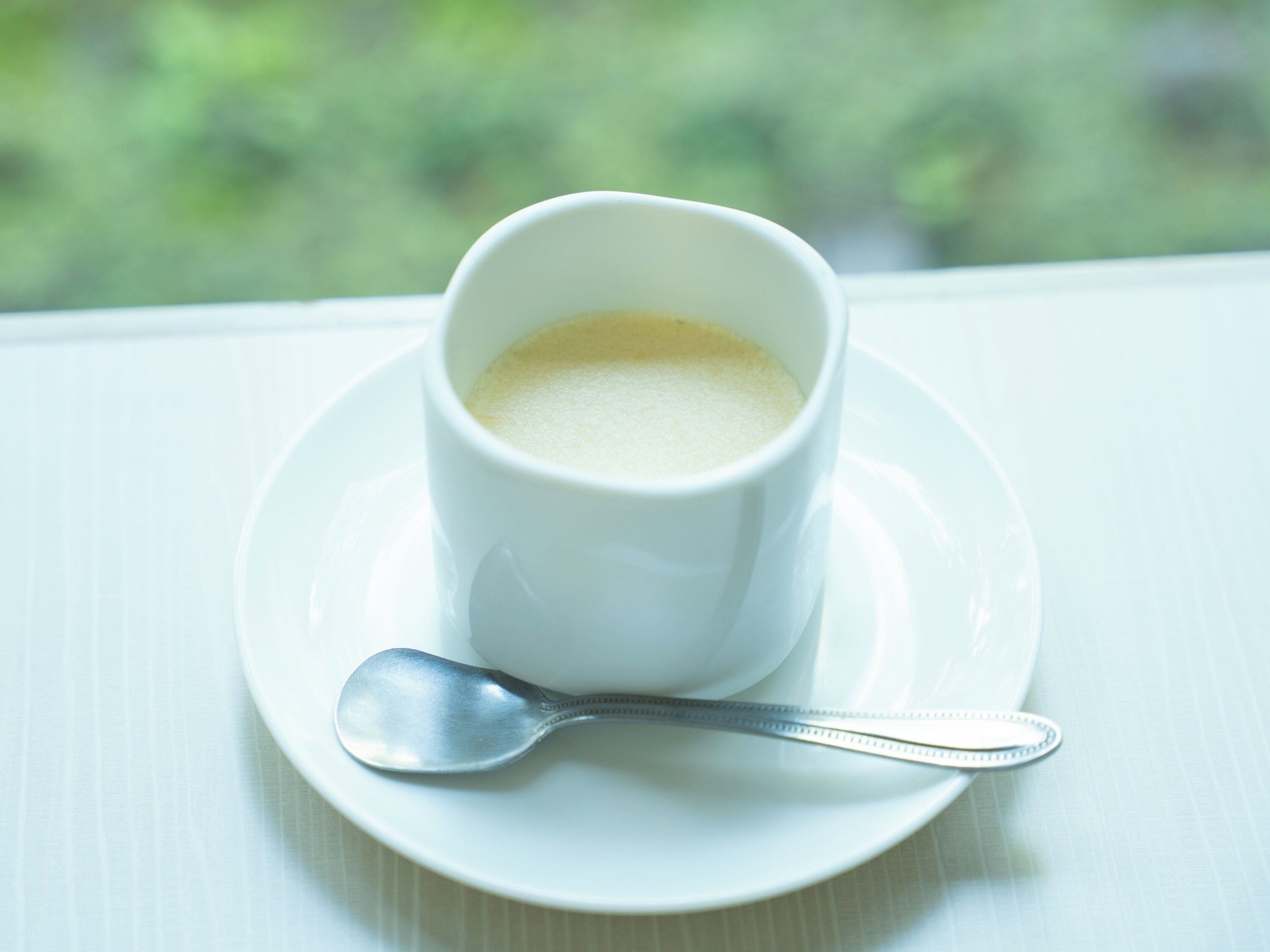A white cup of coffee on a saucer with a spoon beside it, placed on a table by a window.