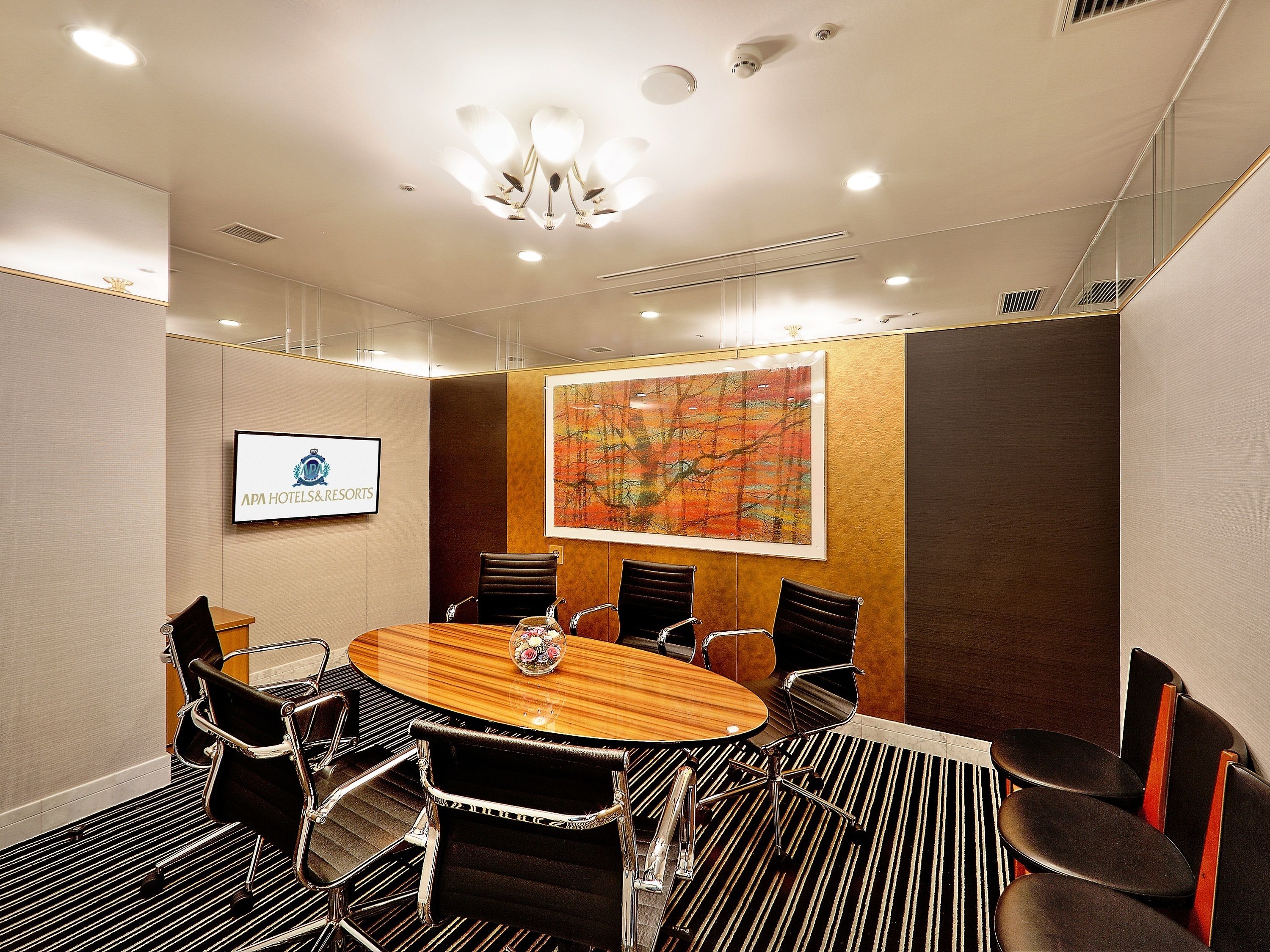 A conference room with a round table, black leather chairs, a chandelier, a framed picture, and a sign for APA Hotels & Resorts.