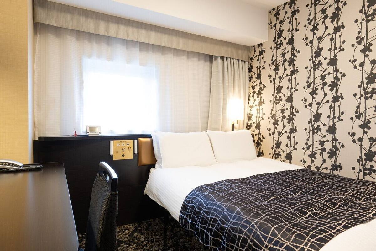 Hotelkamer met een bed, bureau en stoel. Er is een raam met gordijnen, een muur met bloemenpatroon en een wandklok.
