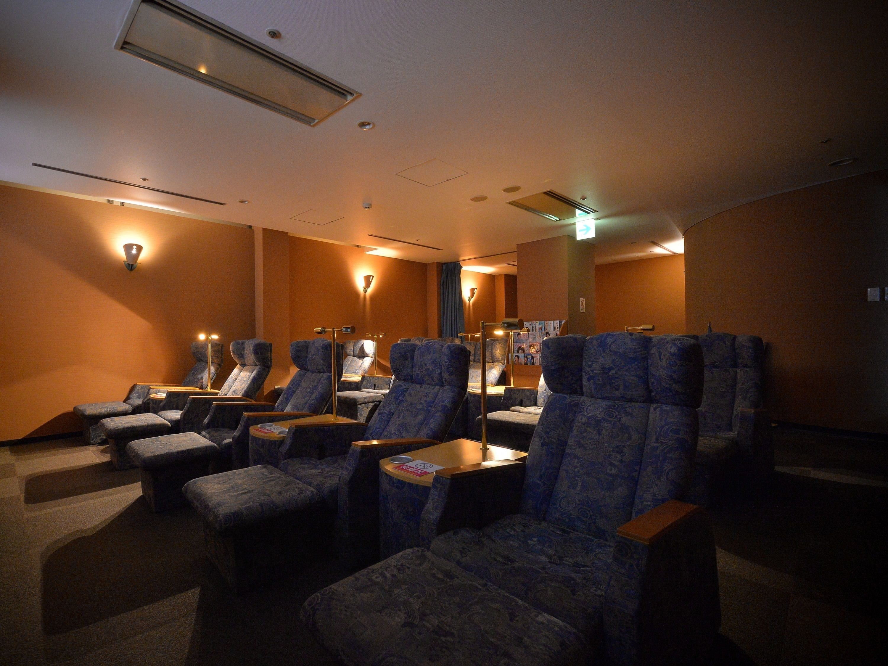 A cinema with rows of blue patterned seats, overhead lighting, and a visible exit sign.