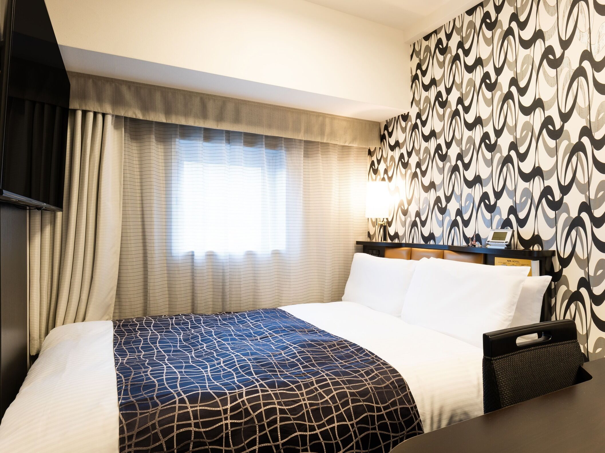 A hotel room with a bed, curtains, a patterned wall, a bedside table with a lamp, and a TV.
