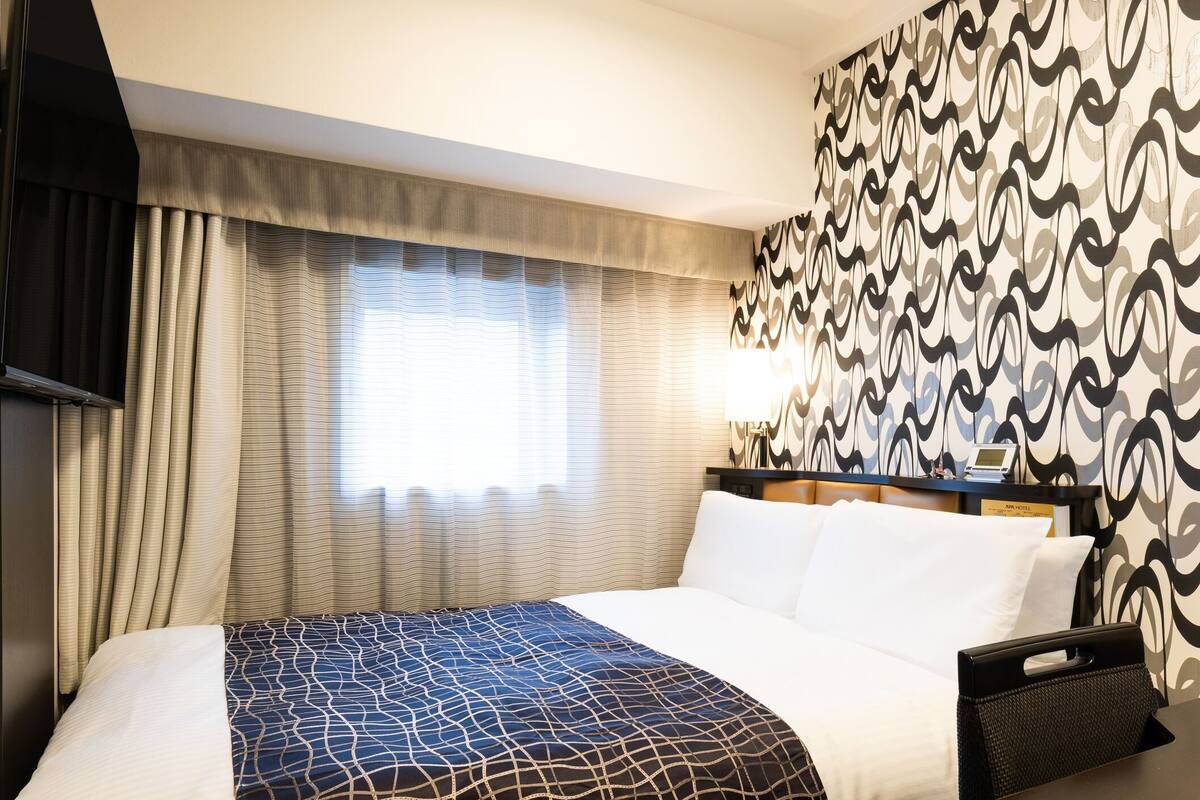 Una habitació d'hotel amb un llit, cortines, un mur amb un disseny i una taula de nit amb una làmpada, i també una televisió.