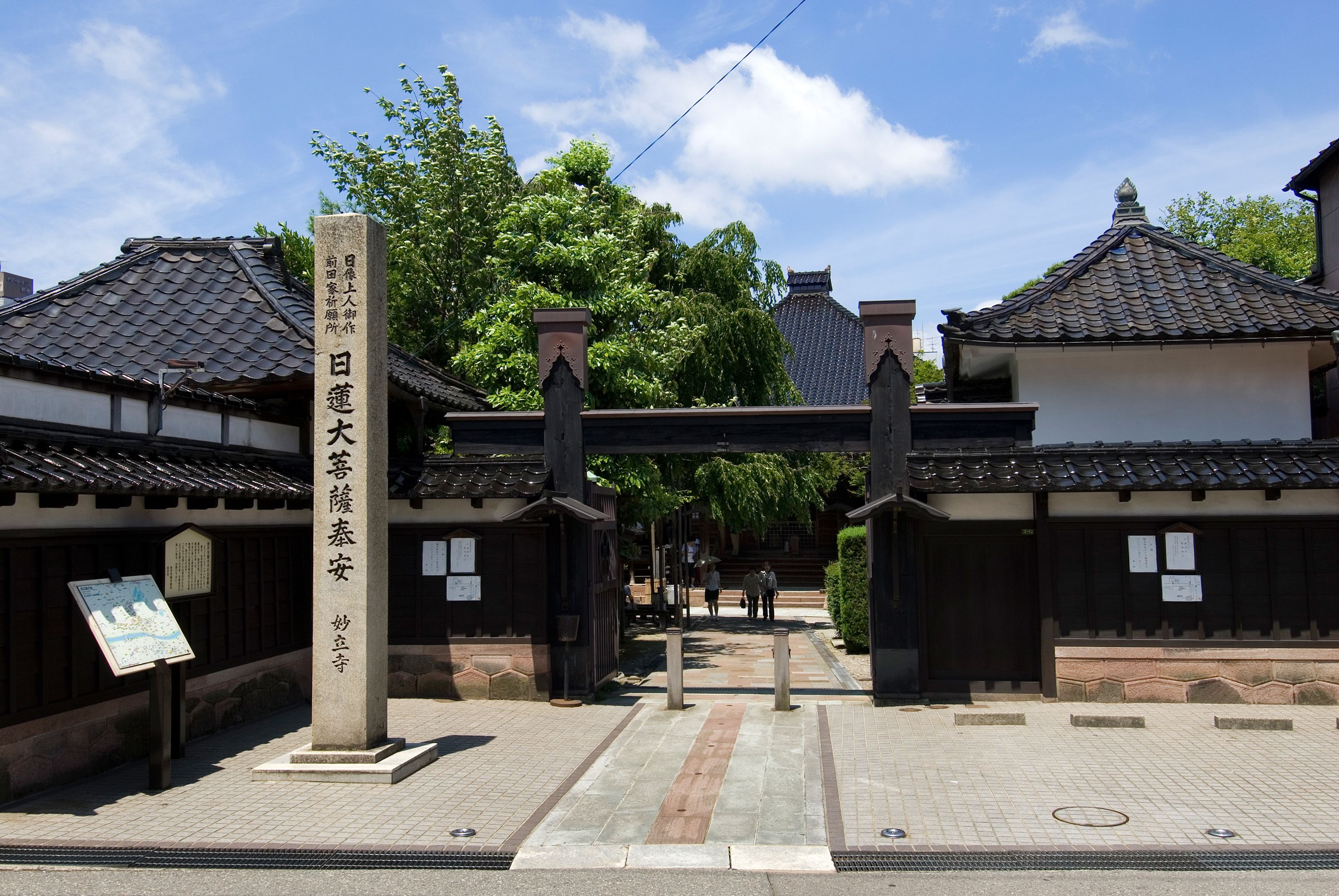A traditional Japanese gate with a stone pillar inscription, leading to a building with tiled roofs and wooden structures.