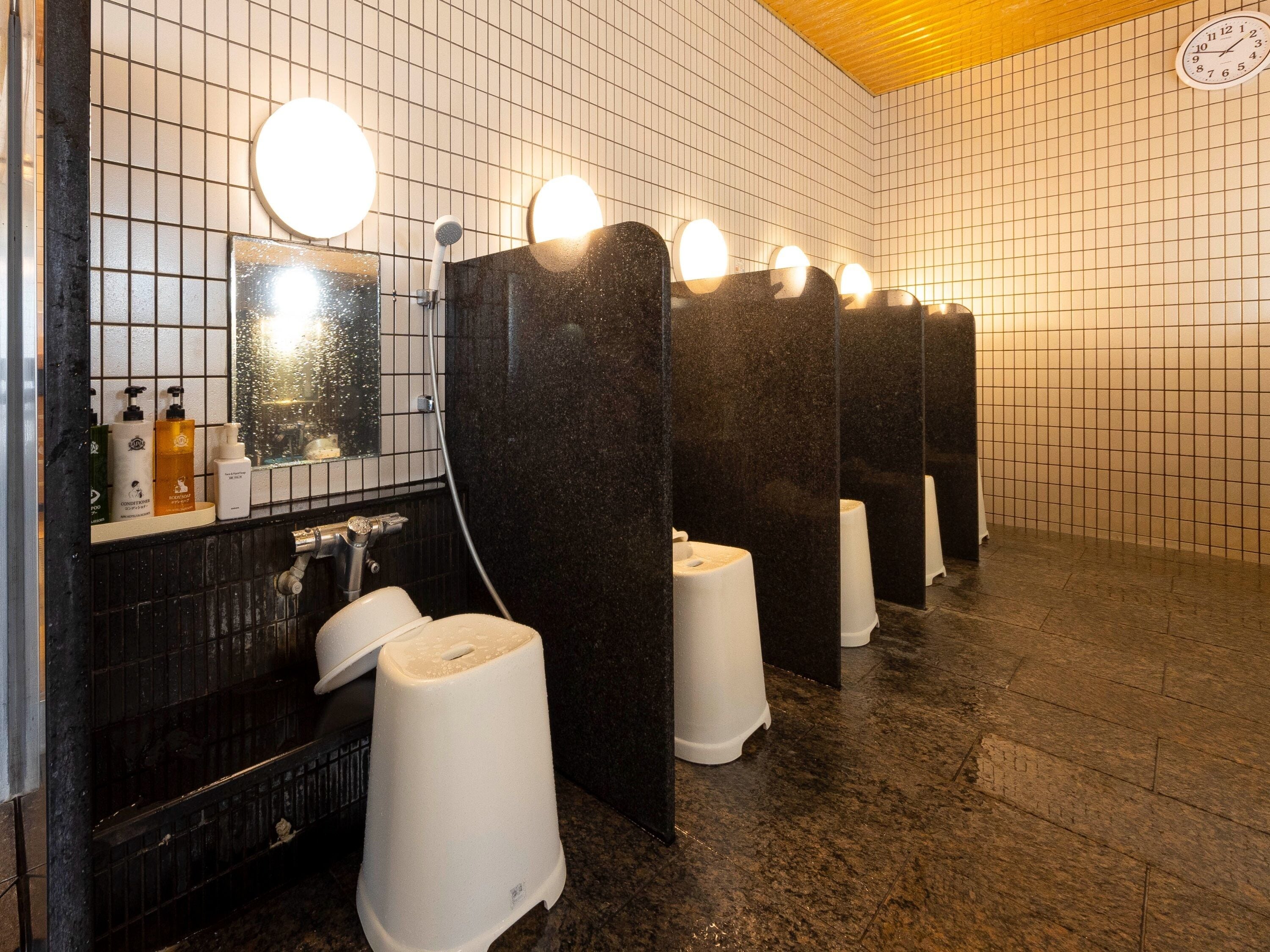 A public restroom with multiple stalls, a mirror, and toiletries on a shelf.
